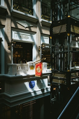 An indoor scene of a mall or shopping center featuring a luxury Ralph Lauren storefront with a Polo brand sign. The architectural design is ornate, with classical elements such as balconies, decorative light fixtures, and intricate moldings. Gold and black decorative motifs adorn the structure in the right side of the image.