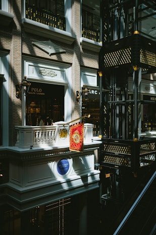 An indoor scene of a mall or shopping center featuring a luxury Ralph Lauren storefront with a Polo brand sign. The architectural design is ornate, with classical elements such as balconies, decorative light fixtures, and intricate moldings. Gold and black decorative motifs adorn the structure in the right side of the image.