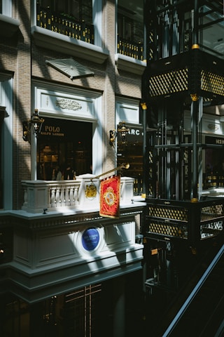 An indoor scene of a mall or shopping center featuring a luxury Ralph Lauren storefront with a Polo brand sign. The architectural design is ornate, with classical elements such as balconies, decorative light fixtures, and intricate moldings. Gold and black decorative motifs adorn the structure in the right side of the image.