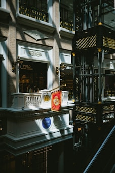 An indoor scene of a mall or shopping center featuring a luxury Ralph Lauren storefront with a Polo brand sign. The architectural design is ornate, with classical elements such as balconies, decorative light fixtures, and intricate moldings. Gold and black decorative motifs adorn the structure in the right side of the image.