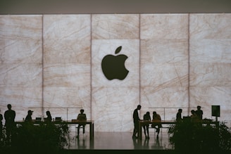 A large Apple logo is prominently displayed on a textured stone backdrop. Silhouettes of people are visible interacting around tables, suggesting a store or lounge setting. The atmosphere is sophisticated and modern.