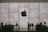 A large Apple logo is prominently displayed on a textured stone backdrop. Silhouettes of people are visible interacting around tables, suggesting a store or lounge setting. The atmosphere is sophisticated and modern.