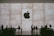 A large Apple logo is prominently displayed on a textured stone backdrop. Silhouettes of people are visible interacting around tables, suggesting a store or lounge setting. The atmosphere is sophisticated and modern.