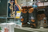 Two blue-coated figurines with large eyes stand inside a clear display case. Behind them, there are other smaller figurines and some packaging, with a depiction of a cartoon dinosaur in the background. The price tag indicating '4,899.00' is visible on the front of the display case.