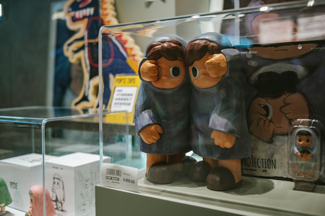 Two blue-coated figurines with large eyes stand inside a clear display case. Behind them, there are other smaller figurines and some packaging, with a depiction of a cartoon dinosaur in the background. The price tag indicating '4,899.00' is visible on the front of the display case.
