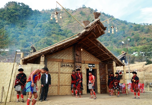 A traditional thatched-roof building made from bamboo stands in a rural setting, with lush green hills in the background. People are dressed in colorful, traditional attire, featuring red, black, and orange patterns, standing and interacting around the building. Some are wearing headgear with feathers and decorative elements. The architecture includes woven bamboo walls and decorative elements hanging from the roof.