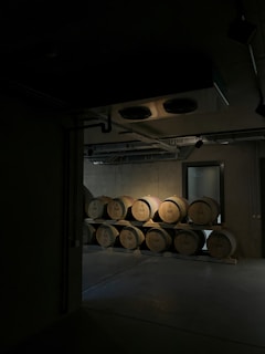 A dimly lit storage room with stacks of wooden barrels organized in two rows. The barrels have some inscriptions on them, possibly for wine storage. The room appears industrial with visible pipes and vents on the ceiling.