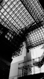 A complex network of intersecting steel and glass structures creates a geometric and modern architectural design. The perspective emphasizes the intricate lattice work against a bright sky, projecting a sense of urban complexity.