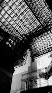 A complex network of intersecting steel and glass structures creates a geometric and modern architectural design. The perspective emphasizes the intricate lattice work against a bright sky, projecting a sense of urban complexity.