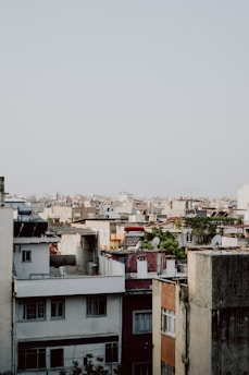 A dense urban landscape features numerous apartment buildings with a variety of rooftops, including solar panels and satellite dishes. The architecture consists of mixed colors and styles, ranging from white to earthy tones. A few trees and patches of greenery are scattered among the buildings.