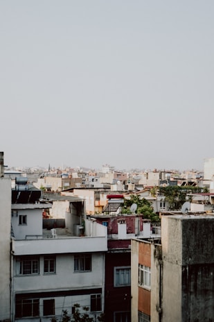 A dense urban landscape features numerous apartment buildings with a variety of rooftops, including solar panels and satellite dishes. The architecture consists of mixed colors and styles, ranging from white to earthy tones. A few trees and patches of greenery are scattered among the buildings.