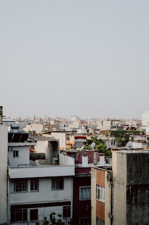 A dense urban landscape features numerous apartment buildings with a variety of rooftops, including solar panels and satellite dishes. The architecture consists of mixed colors and styles, ranging from white to earthy tones. A few trees and patches of greenery are scattered among the buildings.