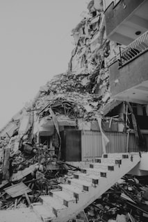 A heavily damaged building with collapsed structures and debris scattered around. The image shows broken concrete, tangled metal rods, and remnants of stairs, indicating significant destruction.