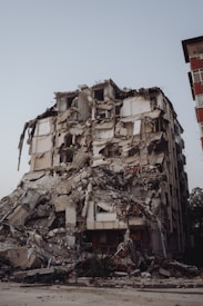 A heavily damaged multi-story building with severe structural collapse, debris scattered around, and visible exposure of internal rooms and materials.