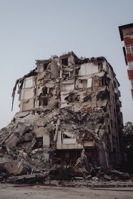 A heavily damaged multi-story building with severe structural collapse, debris scattered around, and visible exposure of internal rooms and materials.