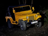 A yellow off-road vehicle with a rugged design is parked on uneven, rocky terrain. The vehicle is illuminated against a dark background, highlighting its sturdy build. The front grille, round headlights, and visible bumper add to its robust appearance. License plate details are visible, but not clearly legible.