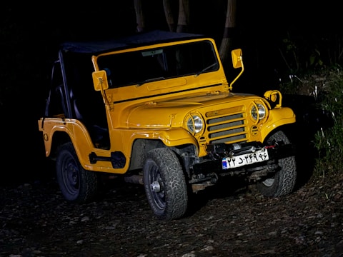 A yellow off-road vehicle with a rugged design is parked on uneven, rocky terrain. The vehicle is illuminated against a dark background, highlighting its sturdy build. The front grille, round headlights, and visible bumper add to its robust appearance. License plate details are visible, but not clearly legible.