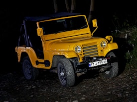 A yellow off-road vehicle with a rugged design is parked on uneven, rocky terrain. The vehicle is illuminated against a dark background, highlighting its sturdy build. The front grille, round headlights, and visible bumper add to its robust appearance. License plate details are visible, but not clearly legible.