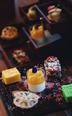 A selection of colorful and intricately arranged desserts is presented on a dark tray. There are layered cakes, a bright yellow roll, a green cube dessert, a creamy dessert topped with orange and a purple edible flower. Surrounding the desserts are pink textured sprinkles.