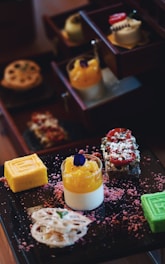 A selection of colorful and intricately arranged desserts is presented on a dark tray. There are layered cakes, a bright yellow roll, a green cube dessert, a creamy dessert topped with orange and a purple edible flower. Surrounding the desserts are pink textured sprinkles.