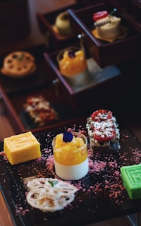 A selection of colorful and intricately arranged desserts is presented on a dark tray. There are layered cakes, a bright yellow roll, a green cube dessert, a creamy dessert topped with orange and a purple edible flower. Surrounding the desserts are pink textured sprinkles.