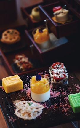 A selection of colorful and intricately arranged desserts is presented on a dark tray. There are layered cakes, a bright yellow roll, a green cube dessert, a creamy dessert topped with orange and a purple edible flower. Surrounding the desserts are pink textured sprinkles.