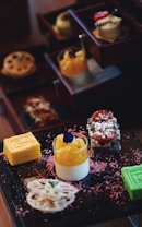 A selection of colorful and intricately arranged desserts is presented on a dark tray. There are layered cakes, a bright yellow roll, a green cube dessert, a creamy dessert topped with orange and a purple edible flower. Surrounding the desserts are pink textured sprinkles.