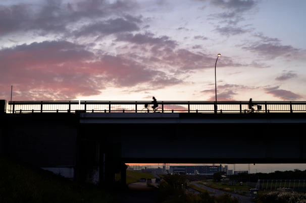 Evening scene of a gravel bike ride event with riders crossing a rustic bridge under warm lights