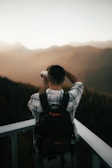 A person wearing a plaid shirt and a backpack is taking a photograph with a camera, surrounded by a vast, hazy landscape of mountains during a sunset or sunrise.