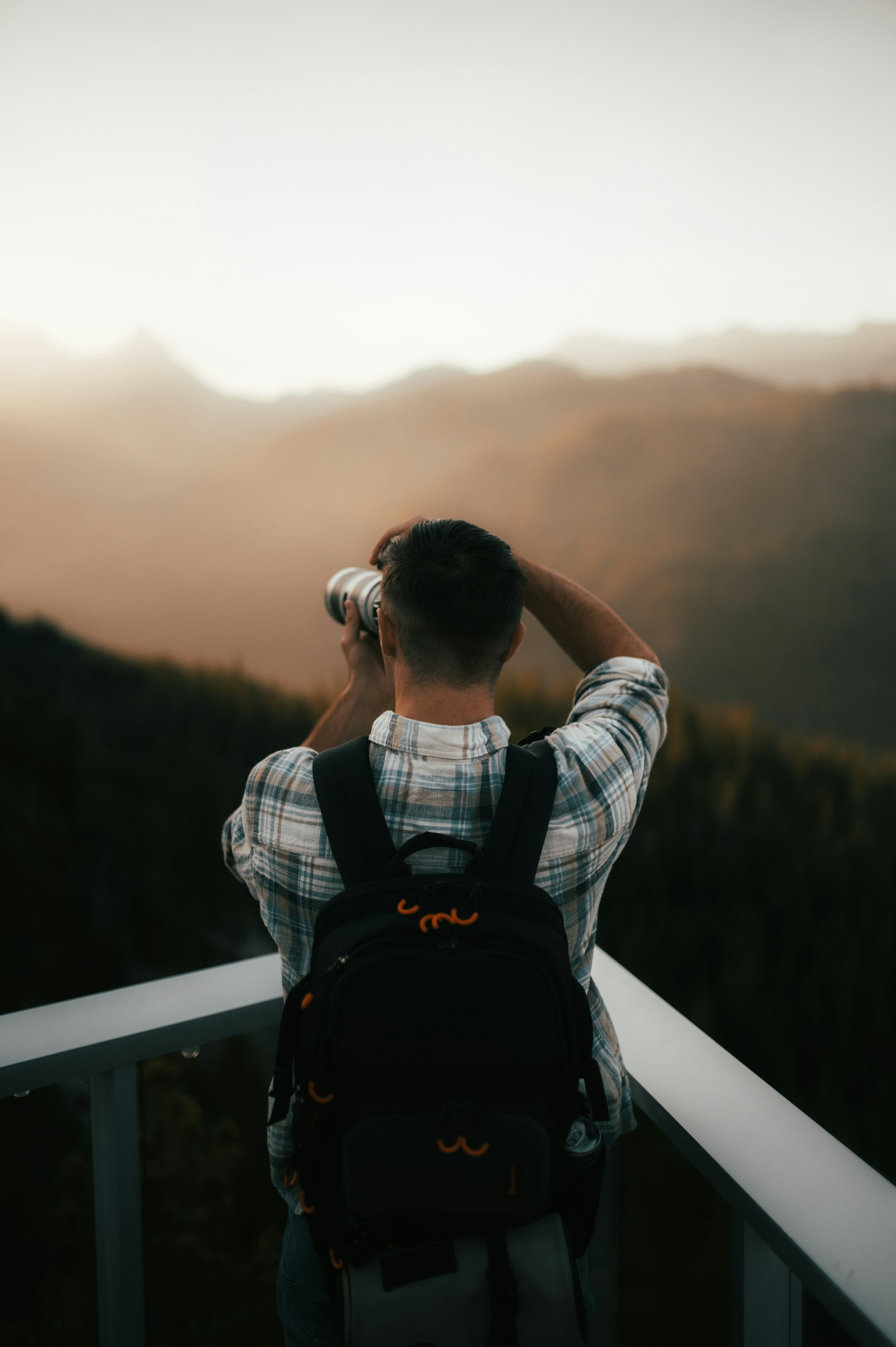 A person wearing a plaid shirt and a backpack is taking a photograph with a camera, surrounded by a vast, hazy landscape of mountains during a sunset or sunrise.