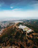 Aerial view of a large observatory situated on a lush, green hill surrounded by dense trees. The landscape gradually transitions into an expansive urban area stretching out to the horizon under a clear blue sky.