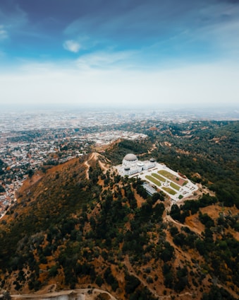 Aerial view of a large observatory situated on a lush, green hill surrounded by dense trees. The landscape gradually transitions into an expansive urban area stretching out to the horizon under a clear blue sky.