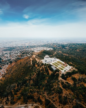 Aerial view of a large observatory situated on a lush, green hill surrounded by dense trees. The landscape gradually transitions into an expansive urban area stretching out to the horizon under a clear blue sky.