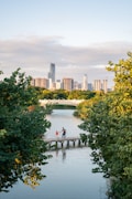 In the foreground, a parent and child are standing on a narrow wooden footbridge over a calm body of water, surrounded by lush green trees. In the background, a city skyline with modern high-rise buildings is visible under a partly cloudy sky, suggesting a park setting within an urban area.