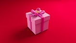 A pink gift box with a large pink ribbon and bow on top, set against a vibrant red background. The box has a yellow decorative center in the middle of the bow.