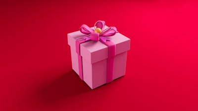 A pink gift box with a large pink ribbon and bow on top, set against a vibrant red background. The box has a yellow decorative center in the middle of the bow.