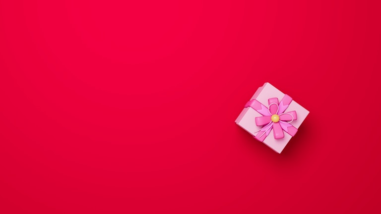 A small, pink gift box adorned with a large bow is placed on a bright red background. The bow is intricately tied and has a yellow center, creating a contrast against the monochrome setting.