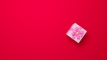 A small, pink gift box adorned with a large bow is placed on a bright red background. The bow is intricately tied and has a yellow center, creating a contrast against the monochrome setting.