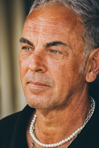 A mature man with gray hair and blue eyes is gazing contemplatively into the distance. His face shows the texture of age, marked by deep lines and wrinkles. He wears a black shirt and is adorned with a pearl necklace.