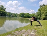 A person is performing a dance or exercise pose near a serene lake surrounded by lush green trees. The scene is calm and the sky is dotted with fluffy clouds, suggesting a pleasant day.