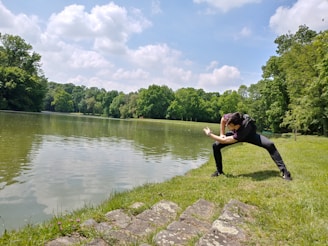 A person is performing a dance or exercise pose near a serene lake surrounded by lush green trees. The scene is calm and the sky is dotted with fluffy clouds, suggesting a pleasant day.
