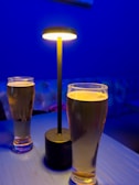 Two tall glasses of beer sit on a blue-lit table, accompanied by a modern, minimalistic table lamp casting a warm glow. The setting suggests an intimate, relaxing atmosphere within a bar or lounge, with a tufted sofa blurred in the background.