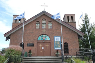 A brick church building features twin bell towers and a cross on the roof. Three flags are flying, and there are decorative plants near the entrance. Stained glass windows adorn the front, with two large wooden doors below displaying gold crosses.