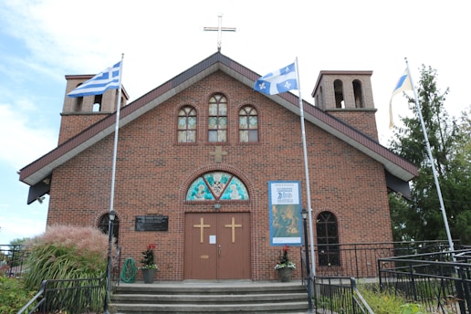 A brick church building features twin bell towers and a cross on the roof. Three flags are flying, and there are decorative plants near the entrance. Stained glass windows adorn the front, with two large wooden doors below displaying gold crosses.