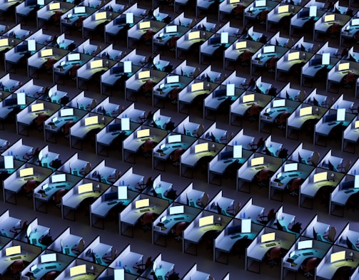 An array of identical office cubicles with computers on each desk, displaying various bright screens. The desks are lined up in a grid pattern, filling the entire space. Each workstation appears to be equipped with standard office items such as chairs, monitors, keyboards, and phones.