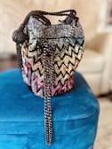 A beaded handbag with a colorful, zigzag pattern resting on a plush, blue surface. The bag is adorned with a long, metal chain strap and features a spherical, beaded accent. The background is softly blurred, drawing focus to the intricate details of the bag.