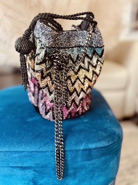 A beaded handbag with a colorful, zigzag pattern resting on a plush, blue surface. The bag is adorned with a long, metal chain strap and features a spherical, beaded accent. The background is softly blurred, drawing focus to the intricate details of the bag.
