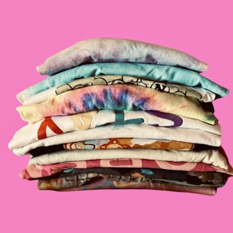 A neatly stacked pile of colorful folded t-shirts is set against a bright pink background. Each shirt features unique patterns and designs, including tie-dye and cartoon motifs.