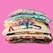A neatly stacked pile of colorful folded t-shirts is set against a bright pink background. Each shirt features unique patterns and designs, including tie-dye and cartoon motifs.