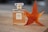 A bottle of perfume labeled 'No. 5 Chamele Paris Eau de Parfum' sits on a wooden surface beside an orange-red autumn leaf. The bottle is clear with a light golden liquid inside, and it has a faceted, clear cap.